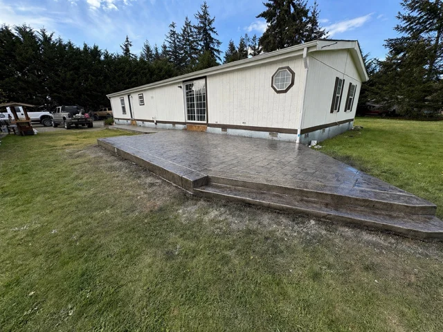 Stamped concrete patio with steps installed beside white mobile home surrounded by evergreen trees